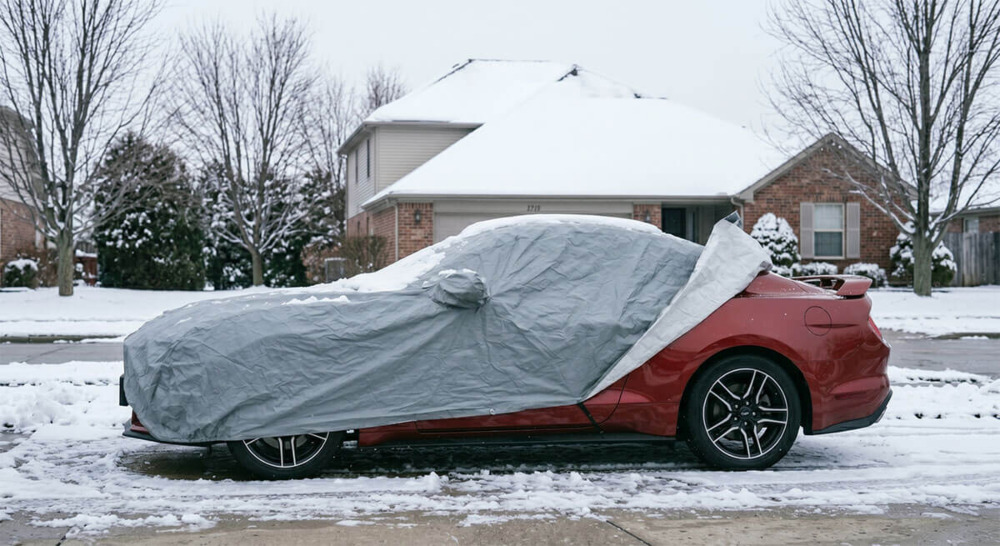 A cheap, ill-fitting gray car cover sagging and slipping off a red Ford Mustang in a snowy New York driveway