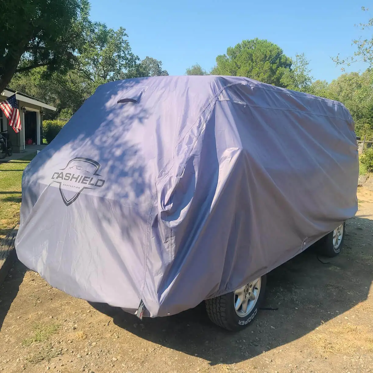 Van under a grey DaShield Ultimum cover in a sunlit yard, front three-quarter view