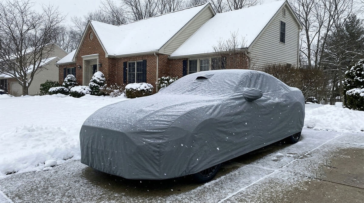 A red Ford Mustang GT fully covered with a gray DaShield car cover in a snowy New York driveway during winter